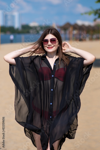 Beautiful, attractive, happy girl posing on a sandy beach on a sunny day in a red swimsuit, sunglasses and a black beach tunic. Beachwear