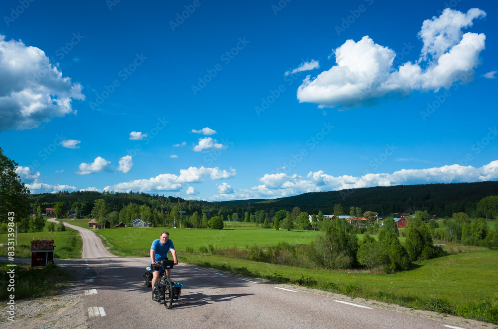 Obraz premium Cycling trip in Sweden, Cyclist on touring bicycle solo travel Nordic countryside, Road among fields summer day, Long distance cycling travel around the world, Mangskogsvägen south of Tobyn, Värmland