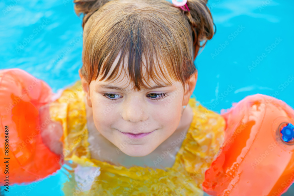 Little preschool girl with protective swimmies playing in outdoor swimming pool by sunset. Child learning to swim in outdoor pool, splashing with water, laughing and having fun. Family vacations.