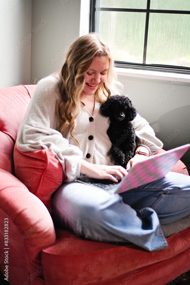 Woman sitting in a pink chair, using a laptop with a black dog o Woman sitting in a pink chair, using a laptop with a black dog o