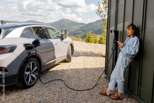 Fotografi Woman stands with a smartphone and waits for electric car to be charged near a charging station at home
