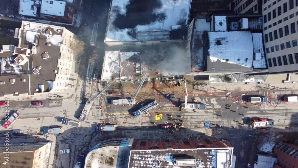 A top down drone shot of black smoke billowing from the rooftop of a building after a devastating fire, on the ground the fired department attempt to contain the smoldering fire A top down drone shot of black smoke billowing from the rooftop of a building after a devastating fire, on the ground the fired department attempt to contain the smoldering fire