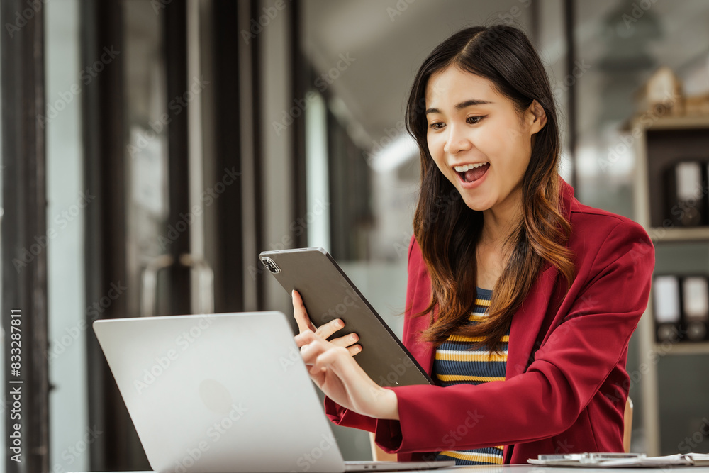 Fototapeta premium A young woman and other Asian individuals in formal suits are seen working at desks with laptops, portraying a successful and happy demeanor.