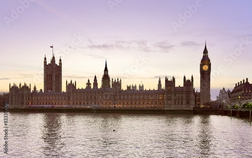 A view of the Houses of Parliament and Big Ben overlooking the River Thames against a purple sky at dusk. 