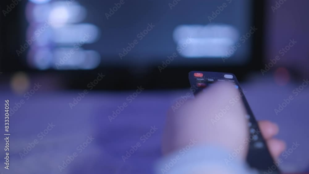 A woman holding a remote control while watching television, with a blurry TV screen in the background. A woman holding a remote control while watching television, with a blurry TV screen in the background.