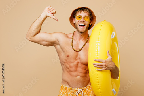 Young sexy man wears yellow shorts swimsuit hat relax rest near hotel pool hold rubber ring point thumb finger on himself isolated on plain beige background. Summer vacation sea sun tan fit concept.