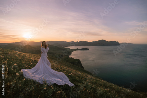 A woman in a white dress stands on a hill overlooking a body of water