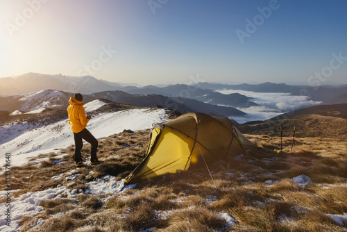 A solo hiker in a yellow jacket stands near a tent on a snowy mountain slope during sunrise  overlooking a breathtaking valley and distant peaks.