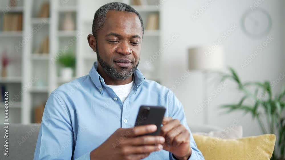 Mature african american man using smartphone sitting on sofa in living room at home. Smiling male browses social networks, does shopping online, chats with a friend, writes or reads messages. Close up Mature african american man using smartphone sitting on sofa in living room at home. Smiling male browses social networks, does shopping online, chats with a friend, writes or reads messages. Close up