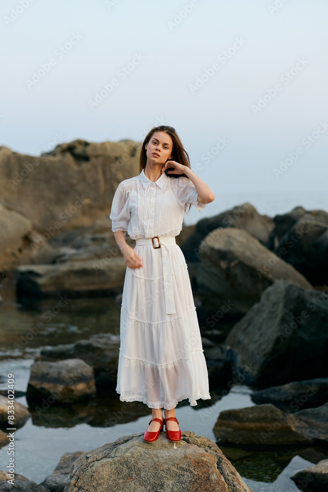 Woman in white dress standing confidently on rocky shore by the ocean Woman in white dress standing confidently on rocky shore by the ocean
