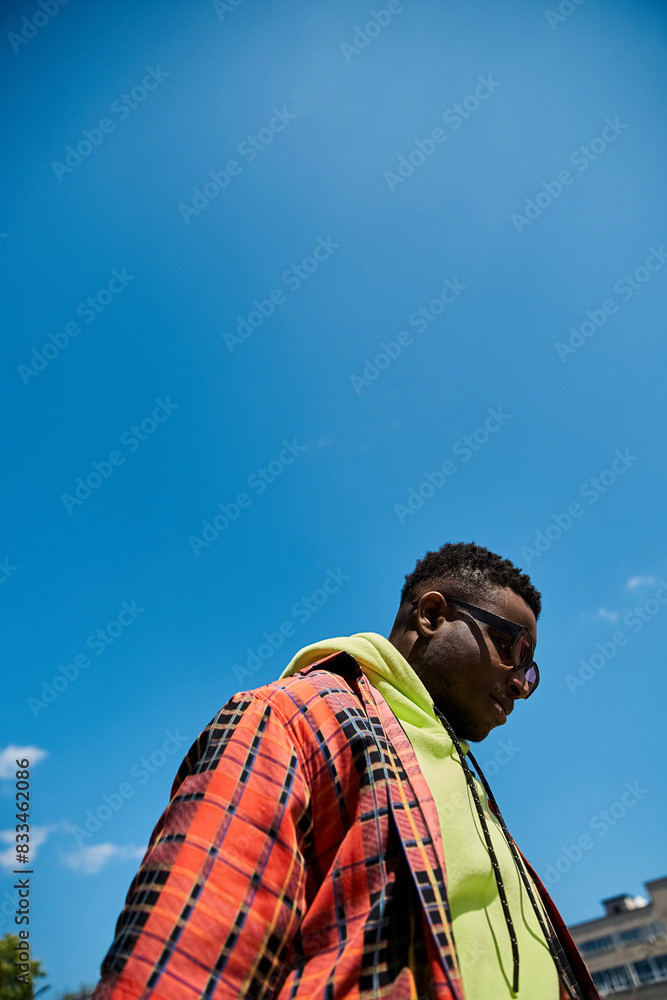 Fototapeta premium Handsome African American man in fashionable plaid jacket standing under a clear blue sky.