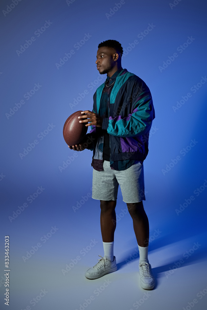 Fototapeta premium A fashionable young African American man holding a football in front of a blue background.