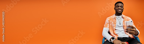 Handsome African American man in fashionable attire sitting elegantly on a chair against an orange background. photo