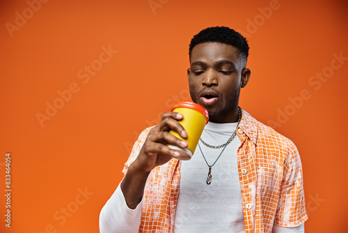 Young African American man drinking from a cup on orange background.