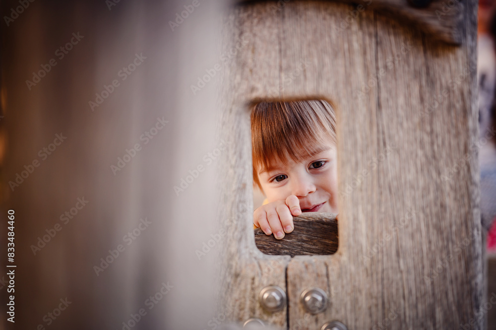 Fototapeta premium A cheerful toddler in overalls peeks through an opening in a wooden playground structure, smiling brightly, with an autumnal background.