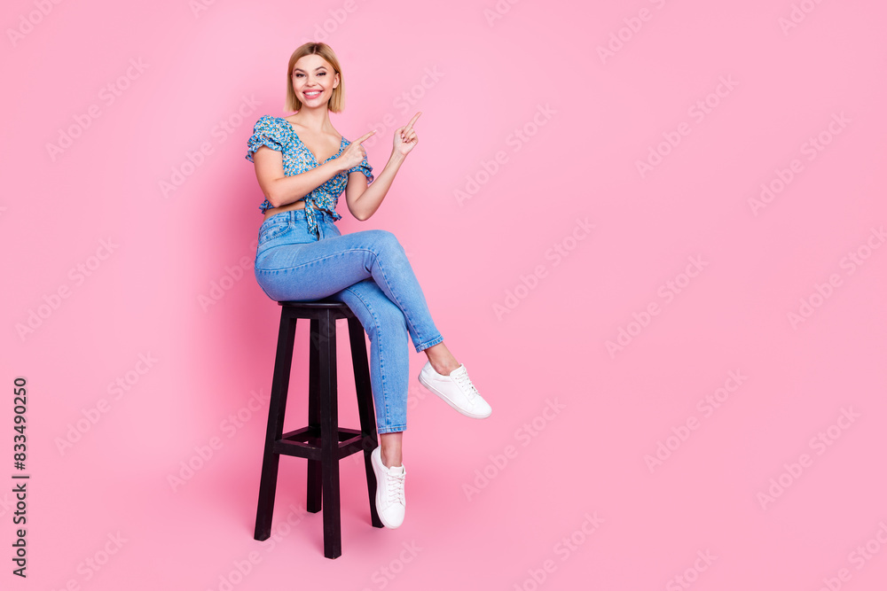 Full size photo of pretty young girl sit stool indicate fingers empty space wear top isolated on pink color background Full size photo of pretty young girl sit stool indicate fingers empty space wear top isolated on pink color background