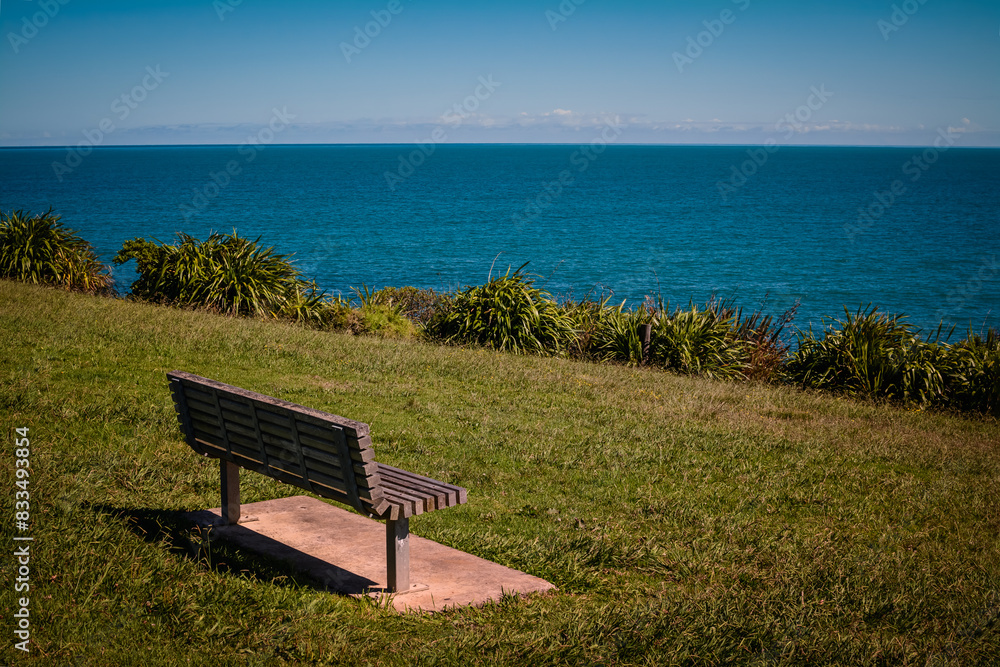A minimalistic landscape with a bench on green grass looking out toward the ocean sea waters. Blue sky and low clouds over distant mountains. Raglan, New Zealand