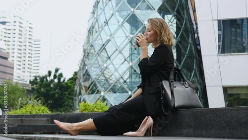 Office worker in metropolis went out for coffee break. Woman took off her shoes, sat down to rest near office on street and drinks coffee. Concept of coffee break from daily office routine.