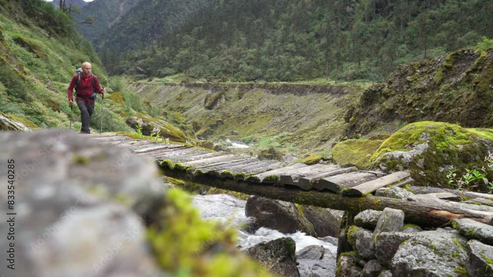 Man with backpack and trekking poles crossing mountain creek wooden bridge during Makalu Barun National Park trek in Nepal. Mountain hiking and active people concept image. Man with backpack and trekking poles crossing mountain creek wooden bridge during Makalu Barun National Park trek in Nepal. Mountain hiking and active people concept image.