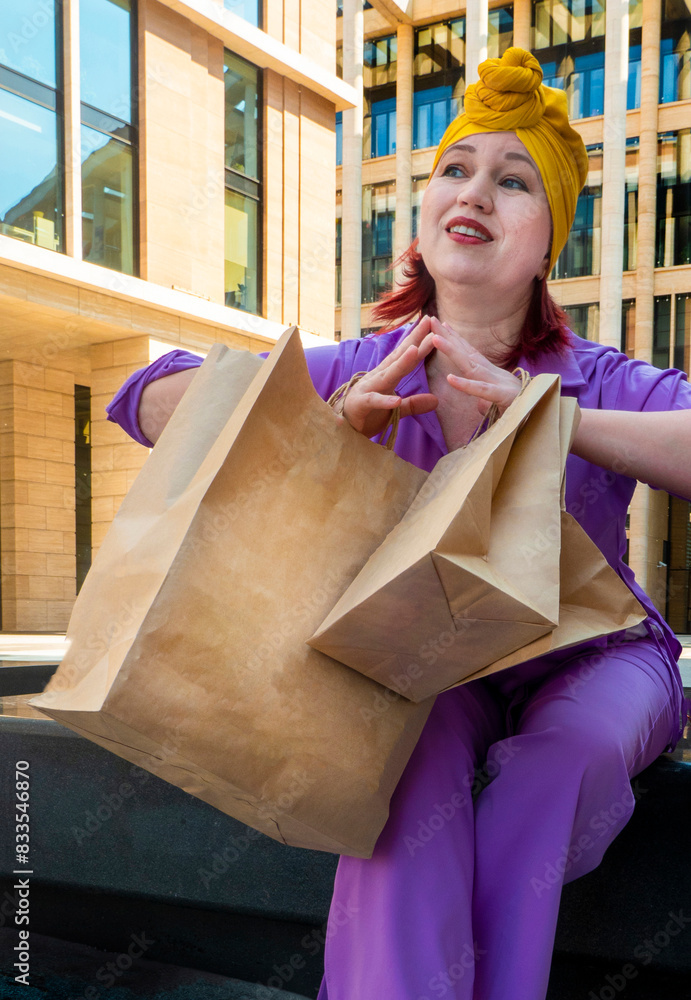 woman with craft shopping bags in the city. The concept of fashionable Consumerism and ecology. An adult Asian woman in a purple suit with red and yellow accents enjoys shopping . a sunny day.