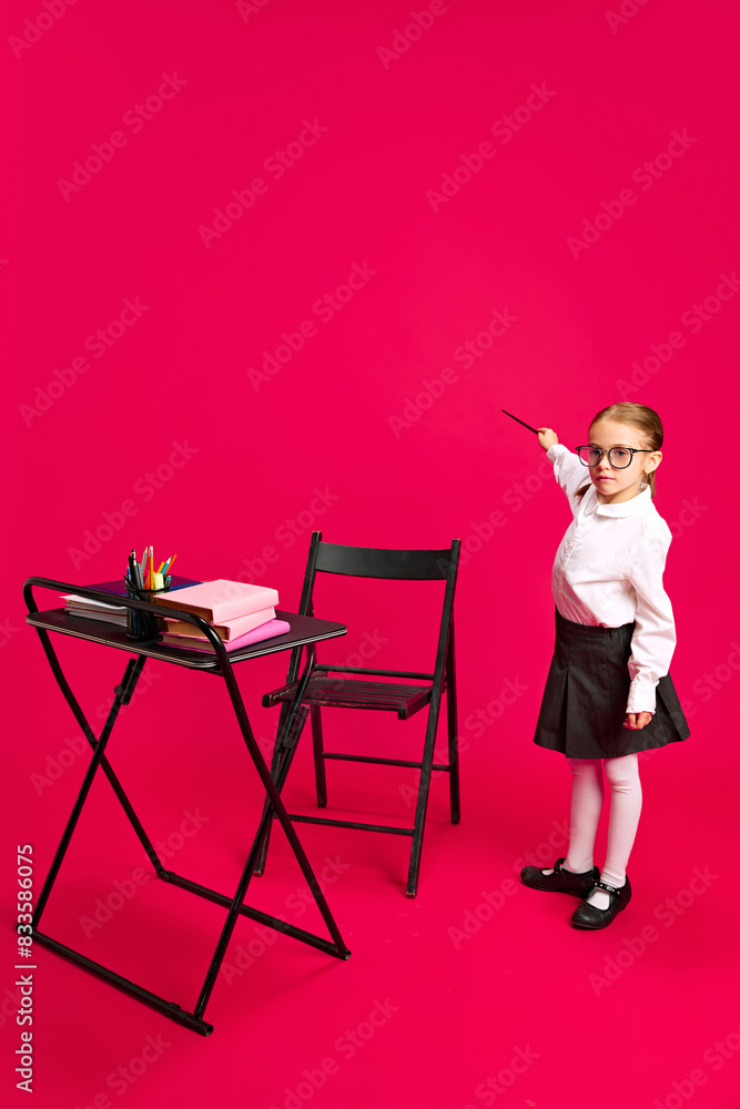Fototapeta premium Child in glasses and formal attire, as teacher holding pointer stick and pointing on imaginative writing board against vibrant pink background. Concept of dreams, future career, games, fantasy. Ad