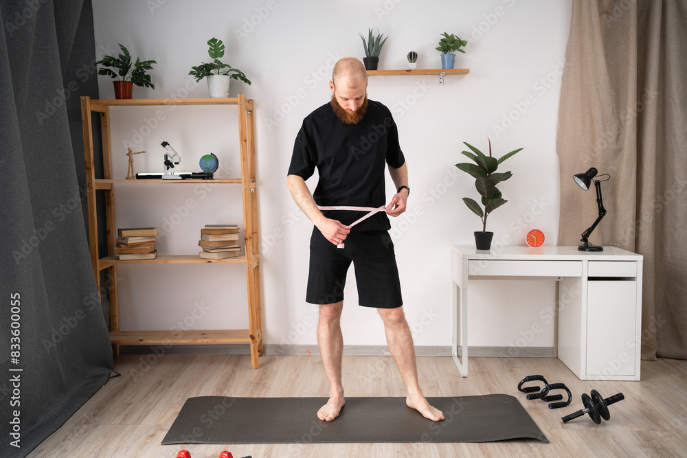 Weight loss and healthy lifestyle concept. Fit fitness man measuring his waistline with measure tape standing on fitness mat. Weight loss and healthy lifestyle concept. Fit fitness man measuring his waistline with measure tape standing on fitness mat.