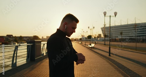 Young active athlete using smartwatch before starting jogging at sunset in the city. Medium slow motion shot from behind. Using wearable technologies in everyday life. Active healthy lifestyle.