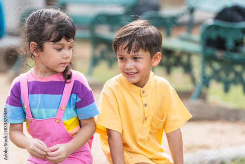 Two of multiracial little children smiling, sitting together outdoors in summer park. Cute kids have fun playing in playground with active activity, holidays and vacations. Selective focus on a boy