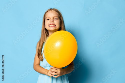 Young girl holding a bright yellow balloon against a vibrant blue backdrop, capturing a moment of joy and color © SHOTPRIME STUDIO