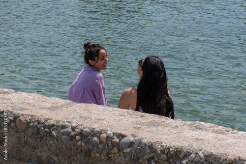 back picture of lesbian couple talking and smiling in an idyllic river landscape, summer evening. On top of a wall their dog