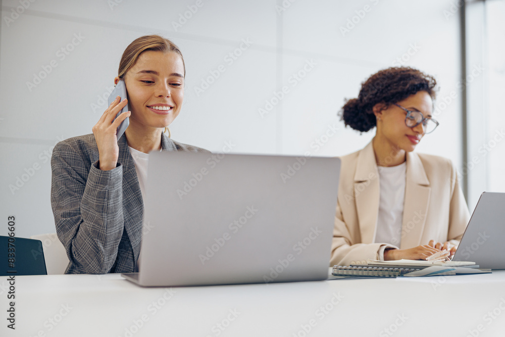 Smiling female sales managers working together in a coworking office while use laptop and smartphone Smiling female sales managers working together in a coworking office while use laptop and smartphone