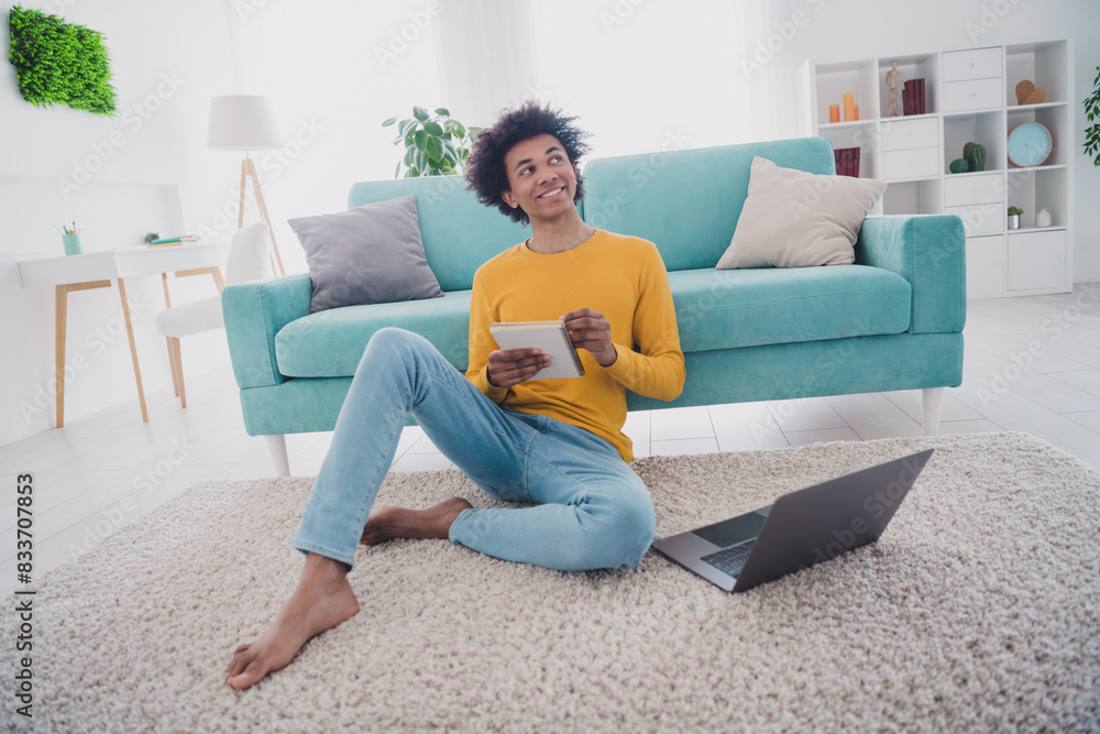 Full size photo of nice young man sit floor hold notepad laptop wear yellow pullover white interior apartment indoors Full size photo of nice young man sit floor hold notepad laptop wear yellow pullover white interior apartment indoors