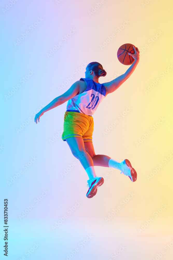Young athletic woman wearing sports uniform doing perfect slam dunk in motion in neon light against gradient studio background. Concept of professional sport, championship, tournament. Ad