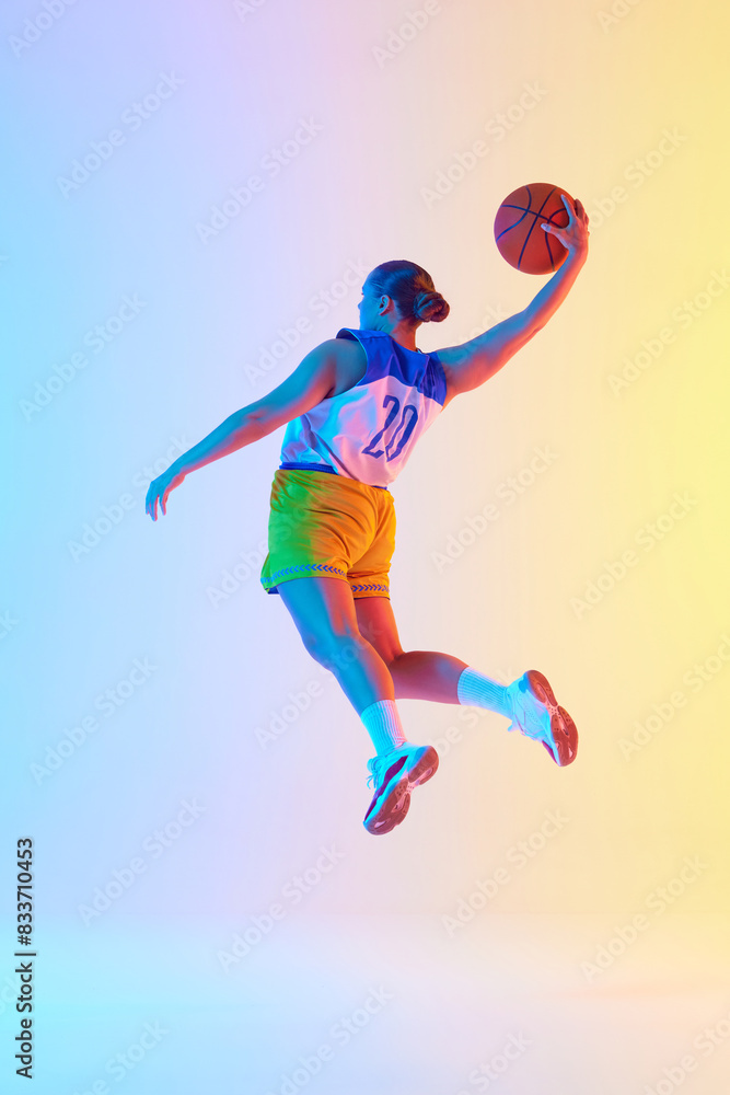 Young athletic woman wearing sports uniform doing perfect slam dunk in motion in neon light against gradient studio background. Concept of professional sport, championship, tournament. Ad Young athletic woman wearing sports uniform doing perfect slam dunk in motion in neon light against gradient studio background. Concept of professional sport, championship, tournament. Ad