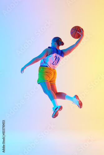 Young athletic woman wearing sports uniform doing perfect slam dunk in motion in neon light against gradient studio background. Concept of professional sport  championship  tournament. Ad