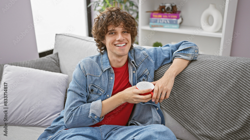 Fototapeta premium A smiling young man with curly hair relaxes on a sofa at home, holding a red mug.