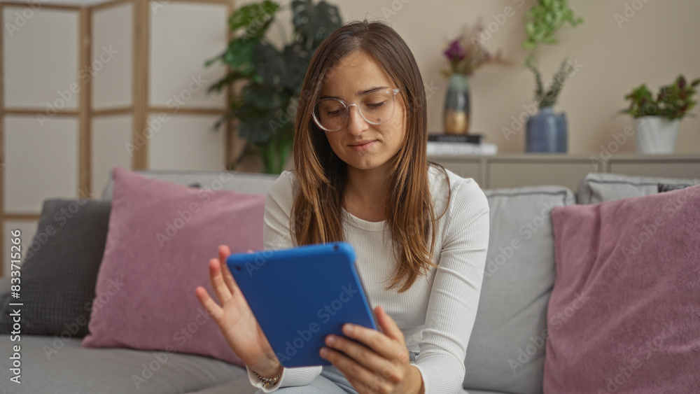 A young, beautiful, blonde woman using a blue tablet while sitting on a grey couch in a cozy living room filled with plants and cushions, showcasing a modern and comfortable home interior. A young, beautiful, blonde woman using a blue tablet while sitting on a grey couch in a cozy living room filled with plants and cushions, showcasing a modern and comfortable home interior.