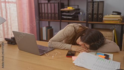 Billede på lærred A young brunette hispanic woman sleeps at her desk in a home office surrounded by work documents, a laptop, and shelves of binders