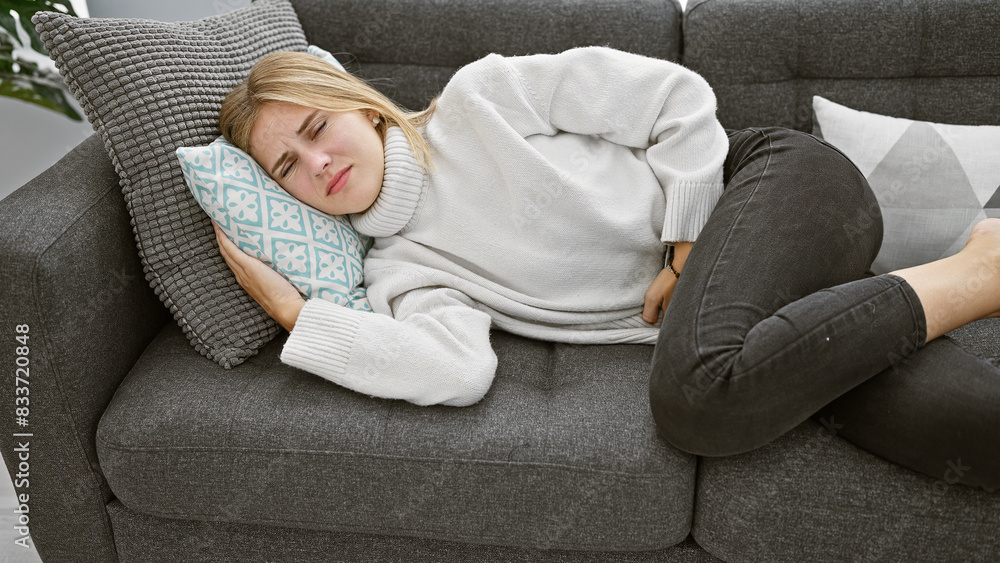 A young blonde woman with blue eyes experiences stomach pain while lying on a couch indoors. A young blonde woman with blue eyes experiences stomach pain while lying on a couch indoors.