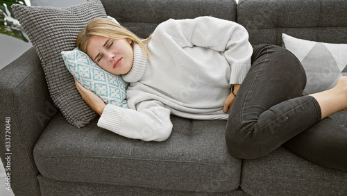 A young blonde woman with blue eyes experiences stomach pain while lying on a couch indoors.