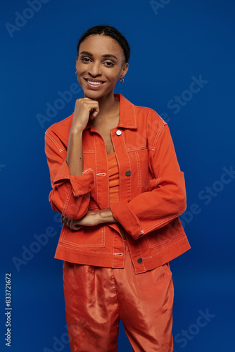 A stylish African American woman flaunts her vibrant orange outfit while striking a pose for a photo on a blue background.