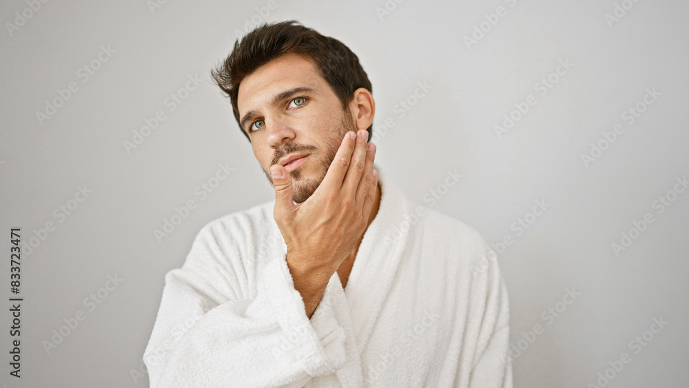 Handsome hispanic man with beard in white bathrobe touches chin against isolated background. Handsome hispanic man with beard in white bathrobe touches chin against isolated background.
