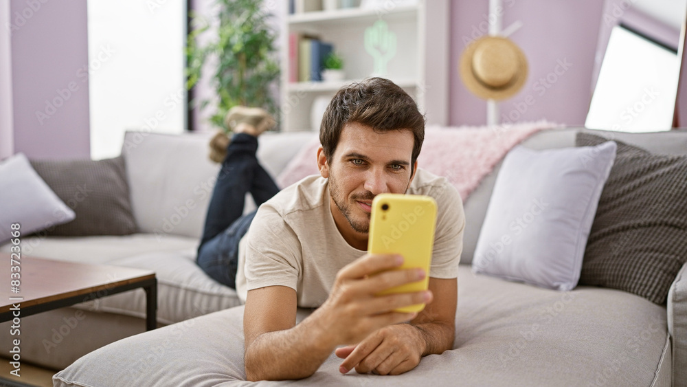 Smiling young hispanic man with a beard using a smartphone in a cozy living room setting. Smiling young hispanic man with a beard using a smartphone in a cozy living room setting.