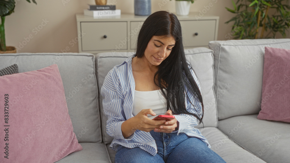 A young hispanic brunette woman is sitting in her living room apartment, using her smartphone on a comfortable gray couch with pink pillows, creating a cozy indoor atmosphere. A young hispanic brunette woman is sitting in her living room apartment, using her smartphone on a comfortable gray couch with pink pillows, creating a cozy indoor atmosphere.