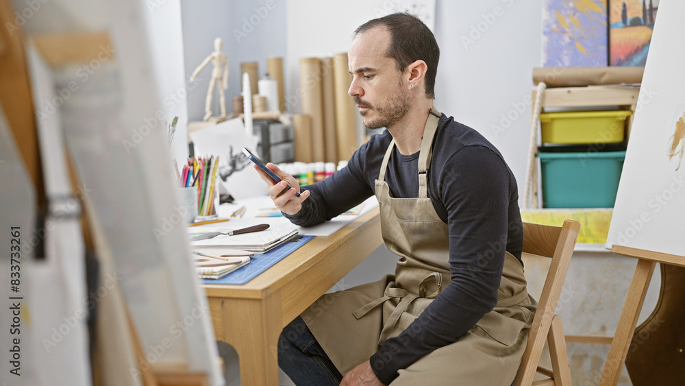 Bald bearded man in studio examining phone with art supplies around. Bald bearded man in studio examining phone with art supplies around.