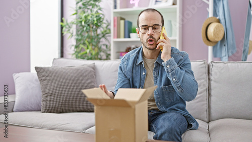 Bald hispanic man with beard talking on phone in a modern living room, perplexed by contents of a carton box. photo