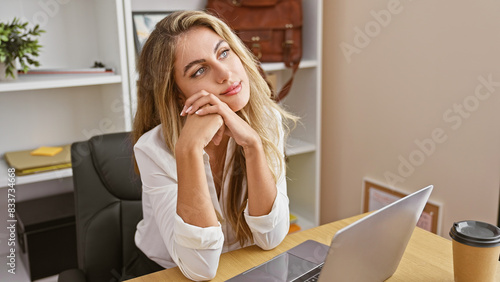 Thoughtful young blonde business worker engrossed in serious work on her laptop in office, portraying the portrait of a successful, beautiful, and professional manager.