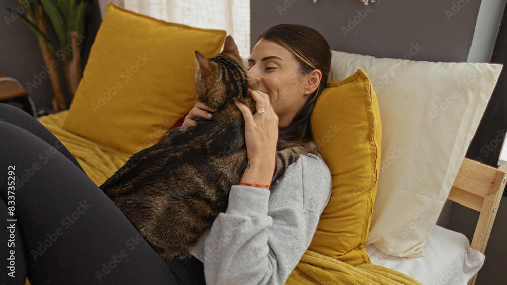 Fototapeta premium A young hispanic woman relaxing in bed at home with a kitten, sharing a tender moment in a cozy bedroom with yellow pillows and soft lighting.