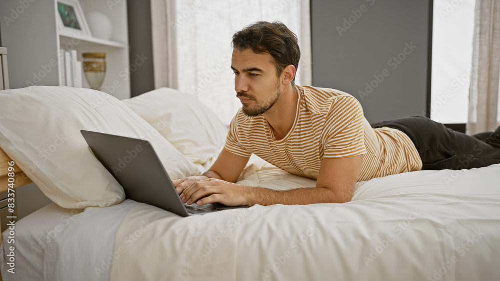 Bearded young hispanic man using a laptop in a modern bedroom setting, exemplifying a casual work-from-home environment. Bearded young hispanic man using a laptop in a modern bedroom setting, exemplifying a casual work-from-home environment.