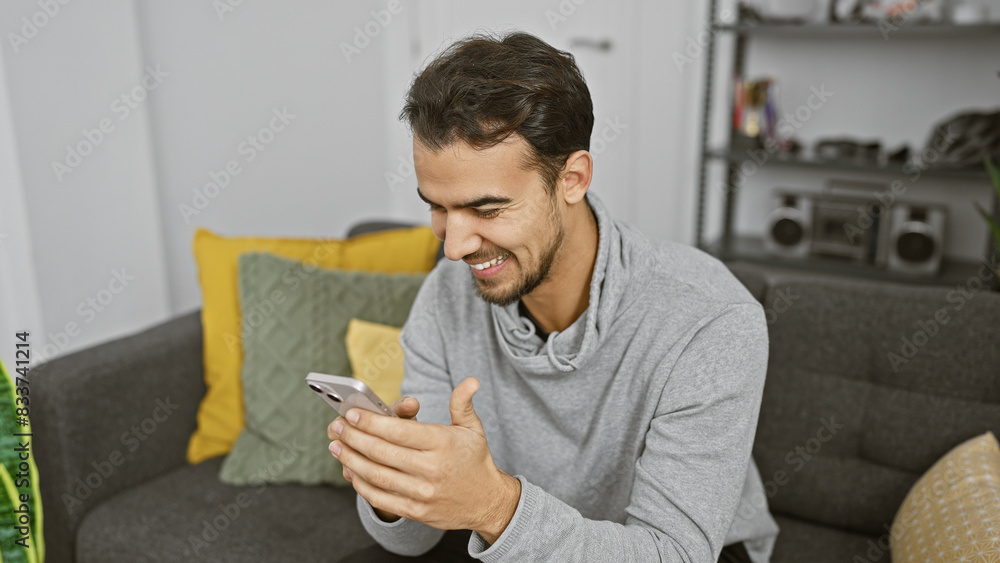 Handsome hispanic man smiling at phone in cozy living room. Handsome hispanic man smiling at phone in cozy living room.